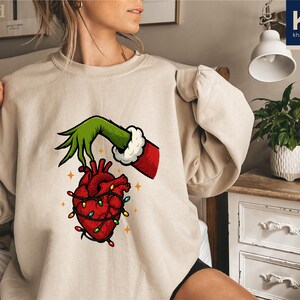 A woman wearing a beige sweater with a graphic of a hand holding a heart with lights around it.
