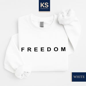 May include: White sweatshirt with the word "FREEDOM" printed in black block letters. The long-sleeved top has a crew neck and a folded sleeve. The brand logo is in the upper left corner, and the color is indicated as "WHITE" in a navy blue rectangle.