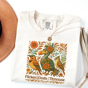 May include: Cream-colored t-shirt featuring a colorful illustration of three birds surrounded by orange flowers and green leaves. The text "Flicker Dodo & Titmouse" is printed at the bottom. A gold necklace is draped over the shirt.