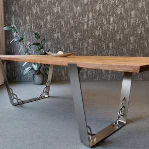 Könnte beinhalten: Ein Esstisch aus Holz mit einer hellbraunen Tischplatte und abgewinkelten, silberfarbenen Metallbeinen. Die Beine haben dekorative Ausschnitte. Eine Pflanze in einem grauen Topf und eine Holzschale sind im Hintergrund zu sehen.