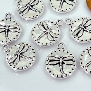 May include: Eight silver-toned metal charms featuring a dragonfly design. Each charm is round and has a textured surface. The charms are perfect for adding a touch of nature to jewelry or crafts.