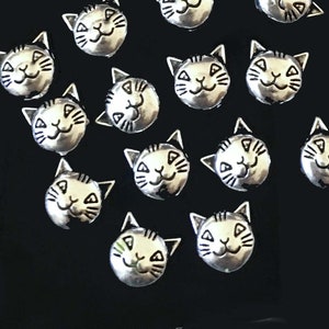 May include: A collection of 14 silver-toned metal charms shaped like cat faces with black eyes and smiling mouths.