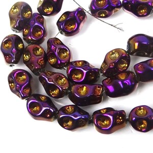 以下が含まれることがあります： 紫と金の頭蓋骨の形をしたビーズのストランド。ビーズはガラス製で、メタリックな仕上げになっています。