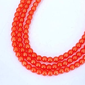 May include: Three strands of orange beads, each composed of small, round beads. The beads are a vibrant, solid orange color. The strands are arranged in a curved, flowing pattern against a white background.