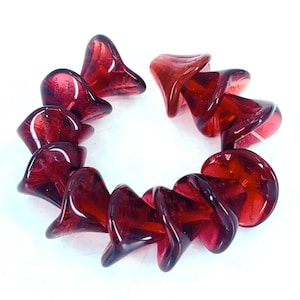 May include: A circular arrangement of deep red glass beads. Each bead has a unique, flared shape, resembling a flower or bell. The beads are translucent, allowing light to pass through, and are set against a white background.