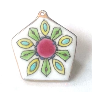 May include: A white, pentagon-shaped pendant with a floral design. The center of the flower is a deep pink, surrounded by green, yellow, and blue petal-like shapes. A small gold loop is at the top for hanging.