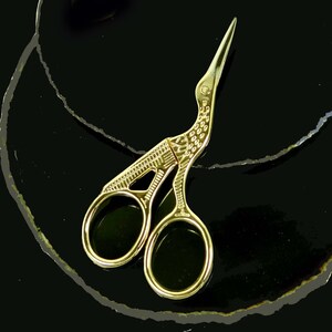 May include: Gold-toned, bird-shaped, decorative scissors with intricate details. The scissors are on a black background.