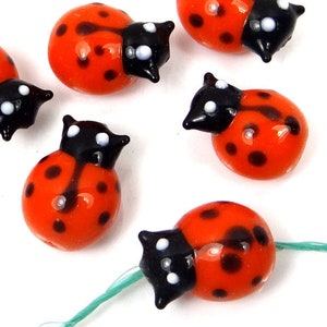 Op de afbeelding: Zes rode en zwarte glazen kralen in de vorm van lieveheersbeestjes met witte stippen. De kralen hebben een diameter van ongeveer 2,5 cm.