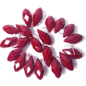May include: A collection of maroon faceted glass teardrop beads. The beads are arranged in a circular pattern, with the points of the teardrops facing outwards.