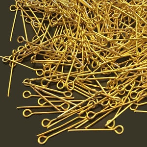 May include: A large pile of gold-colored head pins with small loops at the top. The pins are scattered on a dark brown surface.