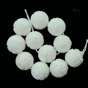 May include: A strand of white, carved beads. Each bead is spherical with a detailed floral or geometric pattern. The beads are strung together on a white string, set against a black background.