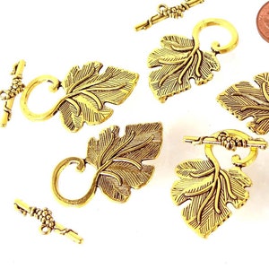 May include: Six gold-tone metal leaf-shaped toggle clasps with a decorative vine design. Each clasp has a loop on one end and a bar on the other end.
