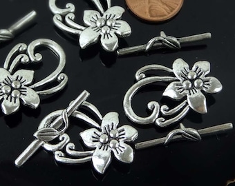 May include: Six silver-toned metal flower toggle clasps with a decorative swirl design. The clasps are approximately the size of a US penny.