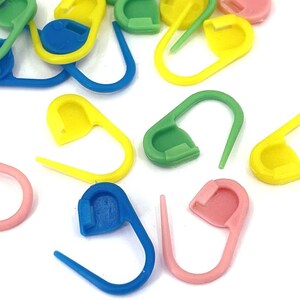 May include: A collection of colorful plastic stitch markers in various shades of pink, yellow, green, and blue. These small, curved markers are used for knitting and crocheting projects. The markers are scattered on a white surface.