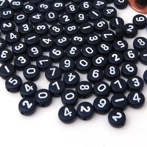 May include: A large quantity of black round beads with white numbers printed on them. The beads are scattered on a white surface.