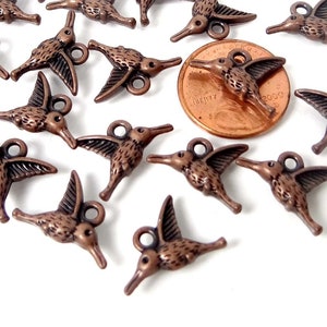 May include: A group of 17 copper-coloured hummingbird charms, each with a loop for attaching to jewellery or crafts. The charms are arranged in a heart shape, with a penny for scale.