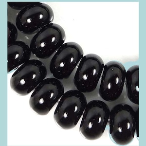 May include: Close-up of a portion of a necklace or bracelet featuring glossy, oval-shaped black beads. The beads are arranged in a curved line against a white background, reflecting light. The jewelry appears to be made of polished stone or glass.