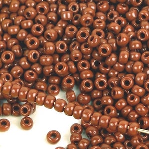 May include: A large pile of brown seed beads. The beads are small and round, with a hole in the center. A strand of the beads is strung on a thread and is lying on top of the pile.