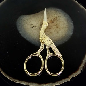 May include: Gold-toned scissors with a bird design. The scissors are on a black and white agate coaster.