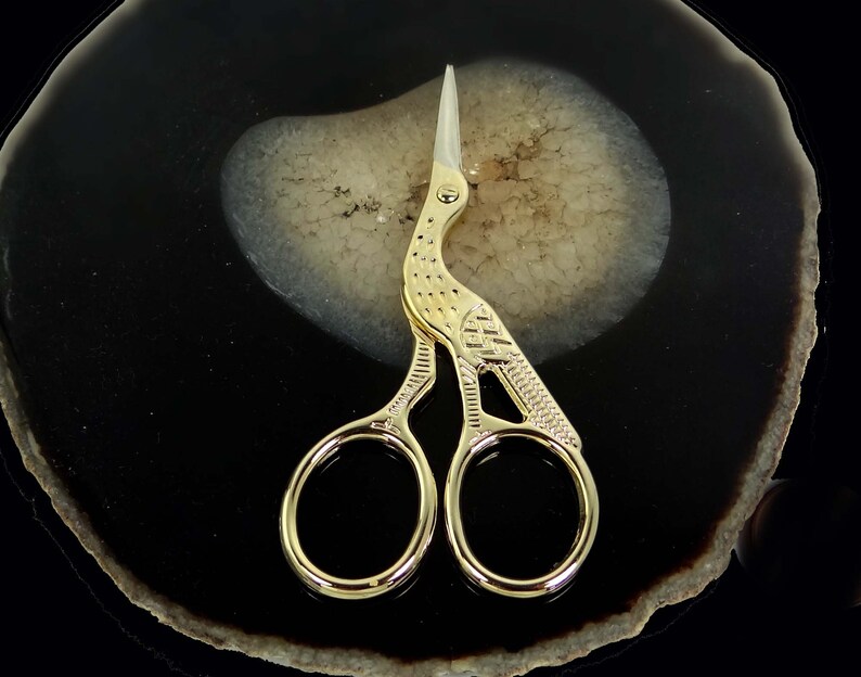 May include: Gold-toned scissors with a bird design. The scissors are on a black and white agate coaster.