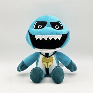 Puede incluir: Un peluche con forma de monstruo de dibujos animados, principalmente azul claro. Tiene una gran boca negra con dientes blancos, ojos negros y un emblema dorado en el pecho.