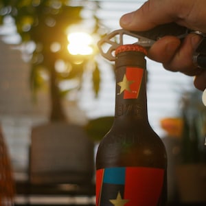 以下が含まれることがあります： 人がマルチツールを使ってビールの瓶を開けています。瓶には星が描かれた赤、白、青のラベルが付いています。マルチツールは赤いキーホルダー付きのキーセットに付いています。