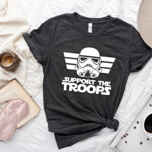 May include: A dark grey t-shirt with a white graphic of a Star Wars Stormtrooper helmet with the text "Support the Troops" underneath.
