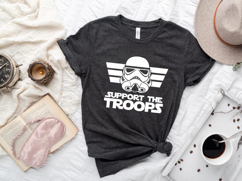 May include: A dark grey t-shirt with a white graphic of a Star Wars Stormtrooper helmet with the text "Support the Troops" underneath.