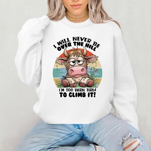 Puede incluir: Sudadera blanca con una vaca de dibujos animados y el texto "I WILL NEVER BE OVER THE HILL I'M TOO DARN TIRED TO CLIMB IT!"