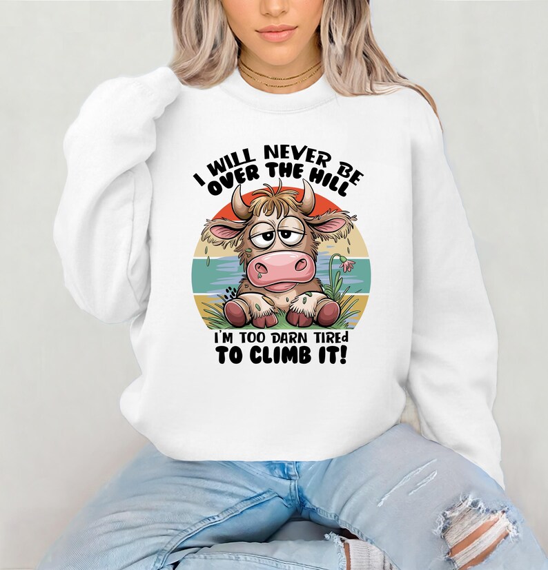 Puede incluir: Sudadera blanca con una vaca de dibujos animados y el texto "I WILL NEVER BE OVER THE HILL I'M TOO DARN TIRED TO CLIMB IT!"