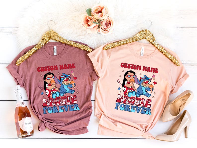 Pu&ograve; includere: Due magliette, una malva e una pesca, ciascuna con una grafica di Lilo e Stitch che mangiano il gelato. Il testo "CUSTOM NAME" e "BESTIE FOREVER" sono stampati sopra e sotto i personaggi.