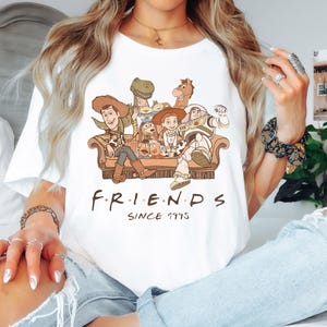 Op de afbeelding: Wit T-shirt met een afbeelding van de Toy Story-personages die op een bank zitten. De tekst "F.R.I.E.N.D.S. SINCE 1995" staat onder de afbeelding.