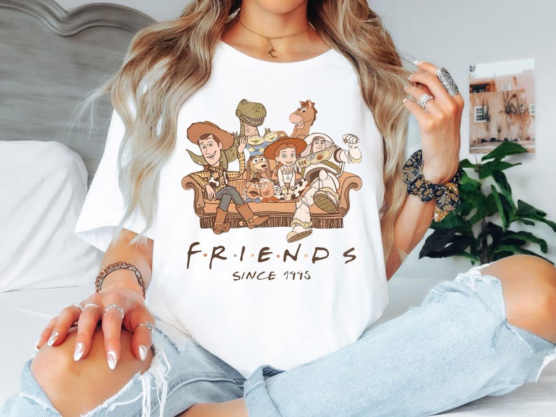 Op de afbeelding: Wit T-shirt met een afbeelding van de Toy Story-personages die op een bank zitten. De tekst "F.R.I.E.N.D.S. SINCE 1995" staat onder de afbeelding.