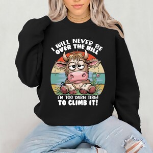 Puede incluir: Sudadera negra con una vaca de dibujos animados y el texto "I will never be over the hill I'm too darn tired to climb it!"