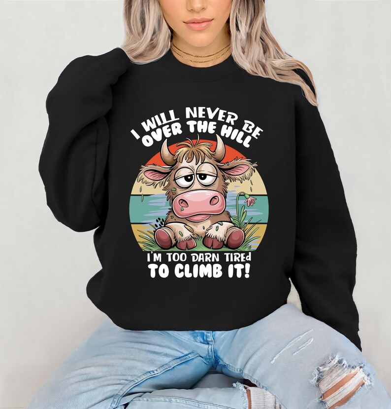 Puede incluir: Sudadera negra con una vaca de dibujos animados y el texto "I will never be over the hill I'm too darn tired to climb it!"
