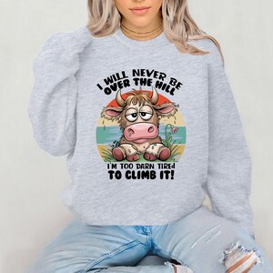 Puede incluir: Una sudadera gris con una vaca de dibujos animados y el texto "I will never be over the hill I'm too darn tired to climb it!"