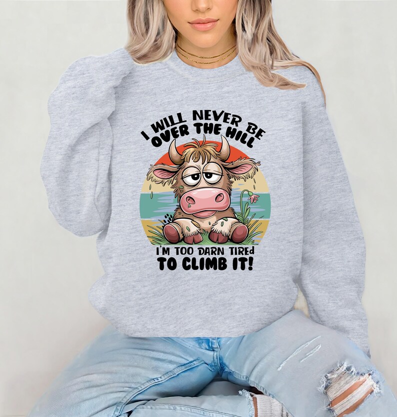 Puede incluir: Una sudadera gris con una vaca de dibujos animados y el texto "I will never be over the hill I'm too darn tired to climb it!"