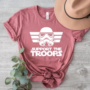 May include: A pink t-shirt featuring a white graphic of a Star Wars Stormtrooper helmet with wings and the text "Support the Troops".