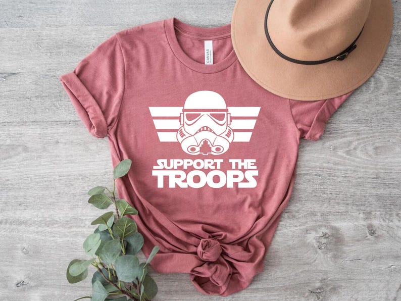 May include: A pink t-shirt featuring a white graphic of a Star Wars Stormtrooper helmet with wings and the text "Support the Troops".