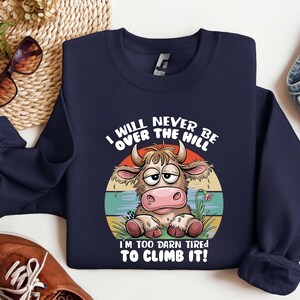 Puede incluir: Una sudadera azul marino con una vaca de dibujos animados con una expresi&oacute;n cansada. El texto en la sudadera dice "I will never be over the hill. I'm too darn tired to climb it!"