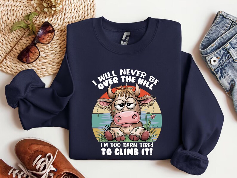 Puede incluir: Una sudadera azul marino con una vaca de dibujos animados con una expresi&oacute;n cansada. El texto en la sudadera dice "I will never be over the hill. I'm too darn tired to climb it!"