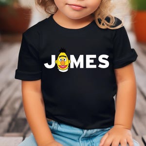 May include: Black t-shirt with the name "JONES" in white block letters and a cartoon character graphic. The graphic is a yellow face with black hair and a red mouth. The shirt is worn by a child.