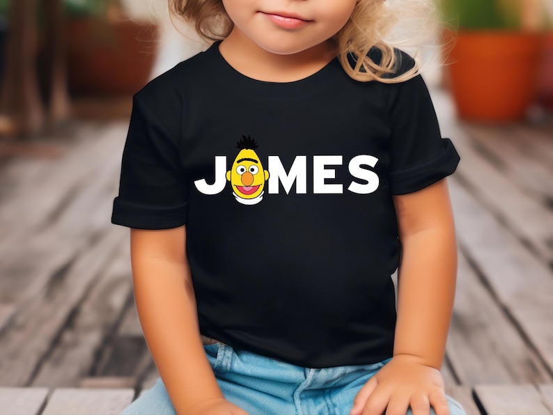 May include: Black t-shirt with the name "JONES" in white block letters and a cartoon character graphic. The graphic is a yellow face with black hair and a red mouth. The shirt is worn by a child.