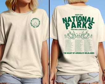 May include: A beige t-shirt with a green graphic on the front that says "The Heart of America's Wildlands". The back of the shirt has a green graphic that says "Keep Our National Parks Pristine & Unspoiled" and lists the names of all the national parks in the United States.