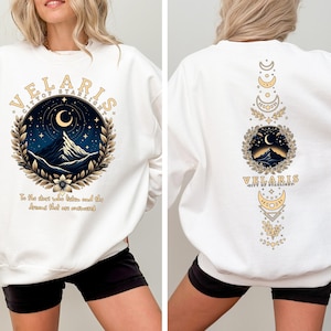 May include: White sweatshirt with a graphic print of a mountain range, moon, and stars in a circle with the text "Velaris City of Starlight" and the text "To the stars who listen and the dreams that are answered". The back of the sweatshirt has a vertical design with a mountain range, moon, and stars in a circle with the text "Velaris City of Starlight" and a series of crescent moons.
