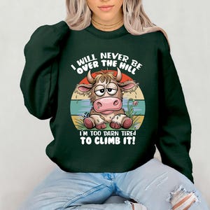 Puede incluir: Sudadera verde con una vaca de dibujos animados y el texto "I will never be over the hill I'm too darn tired to climb it!"