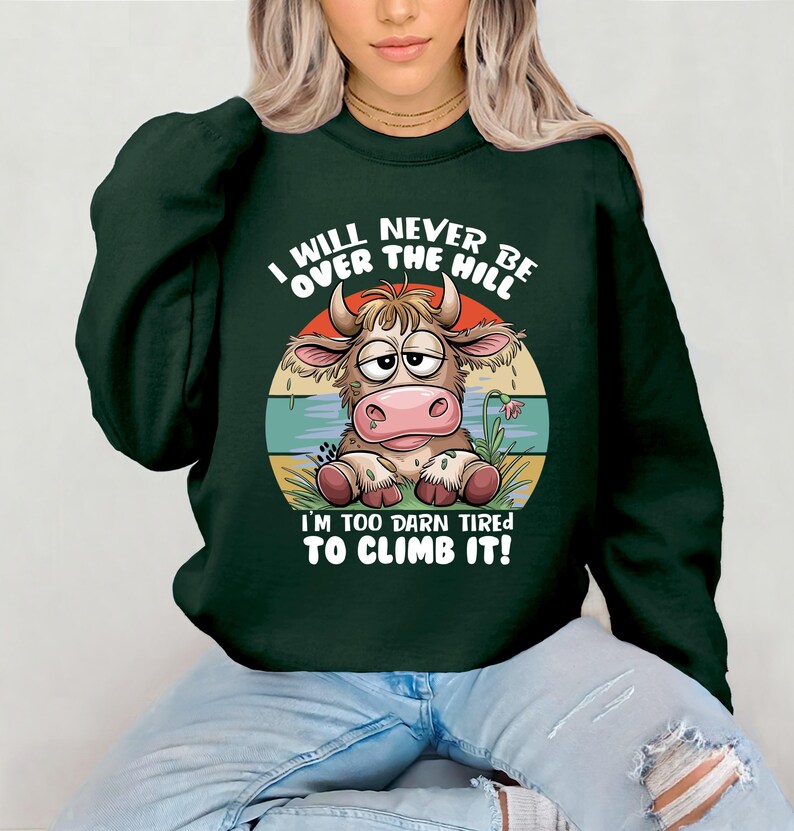 Puede incluir: Sudadera verde con una vaca de dibujos animados y el texto "I will never be over the hill I'm too darn tired to climb it!"