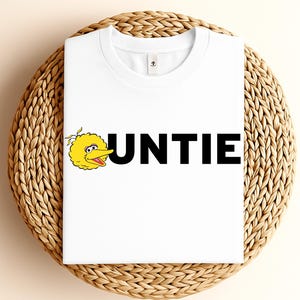 May include: White t-shirt featuring the word "UNTIE" in bold black letters, with a yellow cartoon bird character on the left. The shirt is folded and placed on a woven wicker surface.