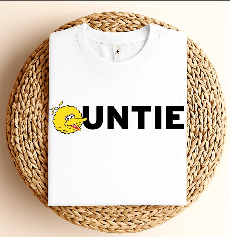 May include: White t-shirt featuring the word "UNTIE" in bold black letters, with a yellow cartoon bird character on the left. The shirt is folded and placed on a woven wicker surface.