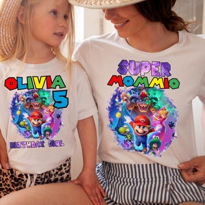 May include: White t-shirts with colorful Mario-themed graphics. One shirt says "Olivia 5 Birthday Girl" and the other says "Super Mommio". The graphics feature Mario, Luigi, and other characters.